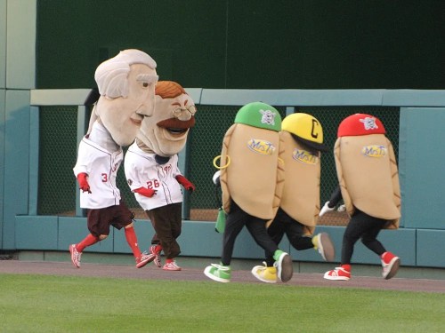 Five costumed mascots, including a giant hot dog and other food characters, race on a baseball field.