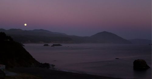 A serene coastal landscape at dusk, featuring a full moon over calm waters and distant mountains.