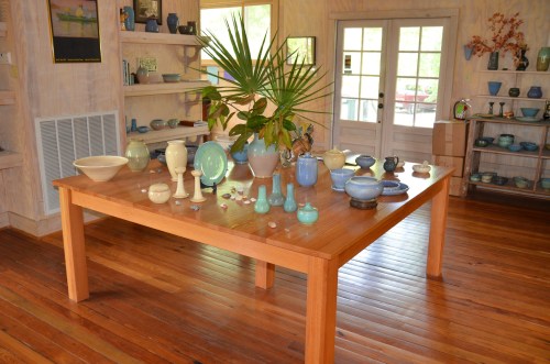 A wooden table displays various ceramic pieces in shades of blue and green, surrounded by a bright, airy room.