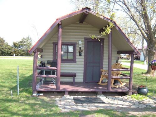 A small, rustic cabin with a sloped roof, porch, and rocking chair, surrounded by green grass and trees.