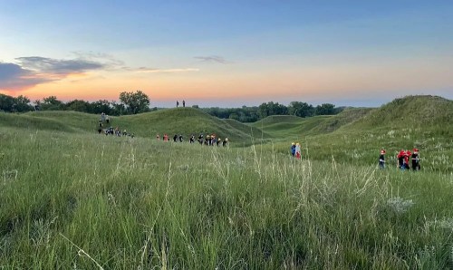 A group of people walking through grassy hills at sunset, with a colorful sky in the background.