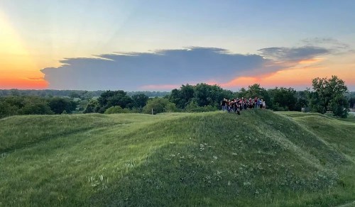 A group of people stands on a grassy hill at sunset, with colorful clouds and a vibrant sky in the background.