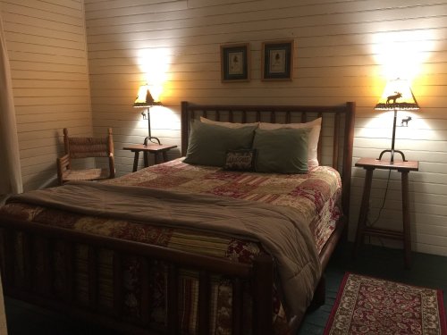Cozy bedroom with a wooden bed, patterned bedding, two lamps, and a chair, featuring warm lighting and a rug.