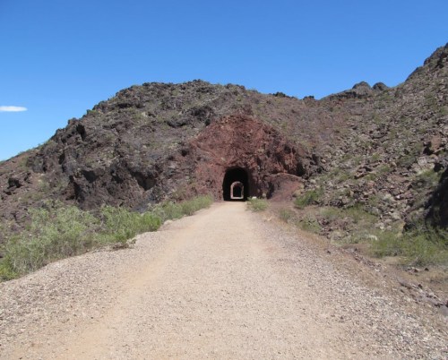 A dirt path leads to a dark tunnel carved into rocky terrain under a clear blue sky.