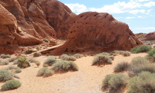 A desert landscape featuring red rock formations and sparse vegetation, with a sign visible in the foreground.