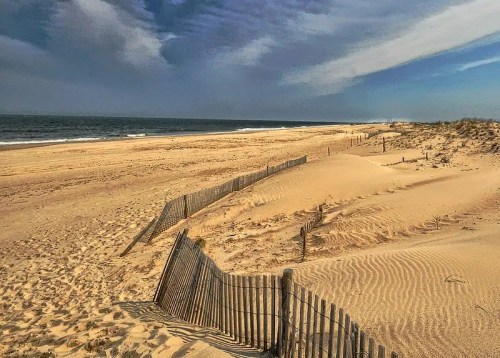 A sandy beach with wooden fencing, gentle dunes, and a calm ocean under a blue sky with scattered clouds.