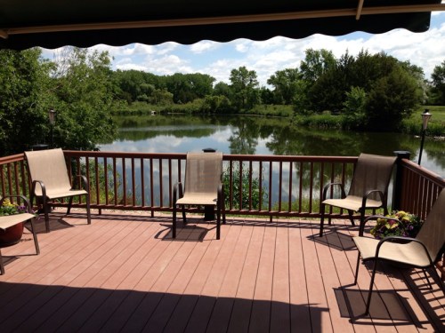 A serene deck with four chairs overlooking a calm lake surrounded by greenery under a blue sky.