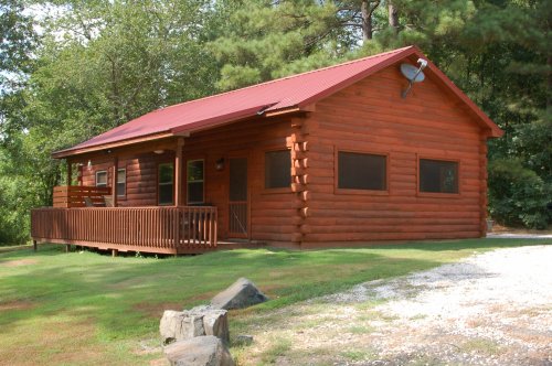 A rustic log cabin with a red metal roof, surrounded by trees and a grassy area, featuring a wooden deck.