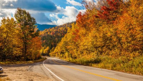 A winding road surrounded by vibrant autumn foliage and mountains under a partly cloudy sky.