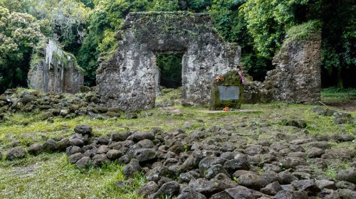 Ruins of an old stone structure surrounded by lush greenery and scattered rocks, with a memorial plaque visible.