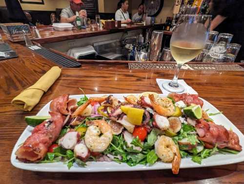 A seafood platter with shrimp, octopus, and prosciutto on a bed of greens, with a glass of white wine in the foreground.