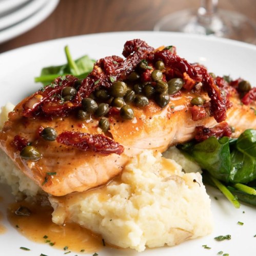 A plate of salmon topped with sun-dried tomatoes and capers, served on a bed of creamy mashed potatoes and spinach.