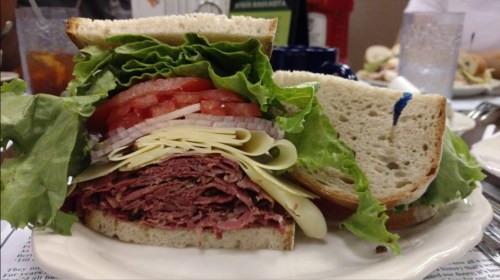 A close-up of a sandwich with layers of lettuce, tomato, cheese, and stacked deli meat on soft bread.