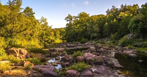 A serene landscape featuring a rocky riverbank surrounded by lush greenery and trees under a clear blue sky.
