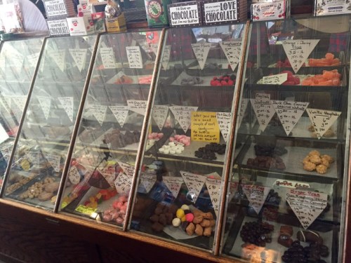 A display case filled with various candies and chocolates, labeled with prices and descriptions on small signs.