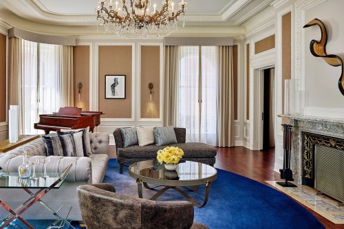 Luxurious living room with a chandelier, elegant furniture, a piano, and a stylish blue rug. Bright and inviting atmosphere.