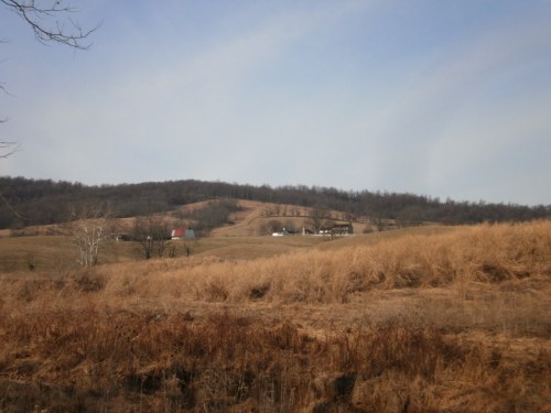 A rural landscape featuring rolling hills, dry grass, and a few distant farmhouses under a cloudy sky.