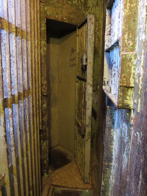 A dimly lit, rusty prison cell with an open barred door and peeling paint on the walls.