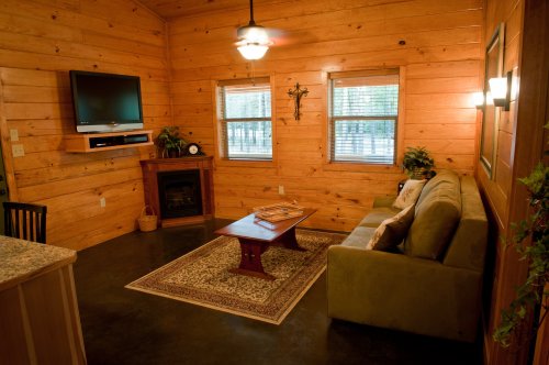 Cozy wooden cabin interior with a sofa, coffee table, TV, and windows, featuring warm lighting and a fireplace.