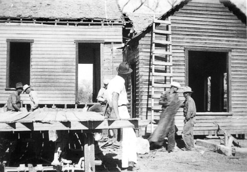 Black and white photo of men working on a wooden house under construction, with ladders and tools visible.