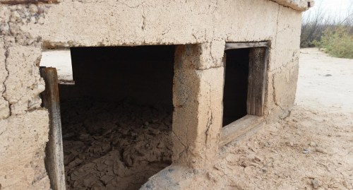 A close-up of a weathered adobe structure with two small windows, showing cracked earth and a sandy landscape.