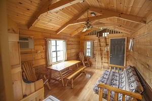 Cozy wooden cabin interior with a dining table, seating area, and a staircase leading to a loft. Natural light from windows.