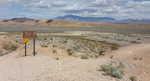 A desert landscape with a signpost indicating a trail, mountains in the background under a cloudy sky.