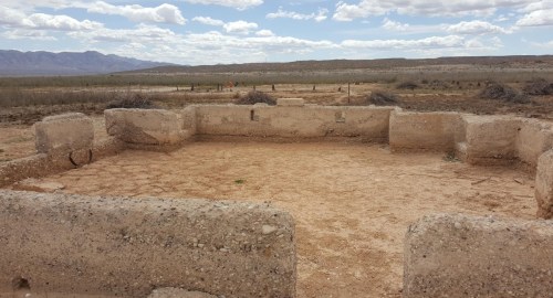 Ruins of a stone structure in a barren landscape, with mountains and clouds in the background.