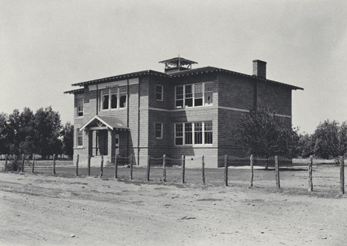 A historic two-story wooden house with a porch, surrounded by a dirt path and sparse trees.