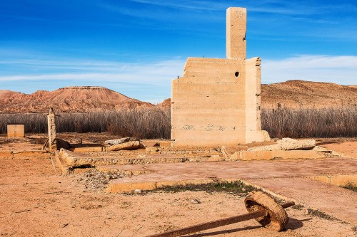 Ruins of a concrete structure in a desert landscape, with mountains and blue sky in the background.