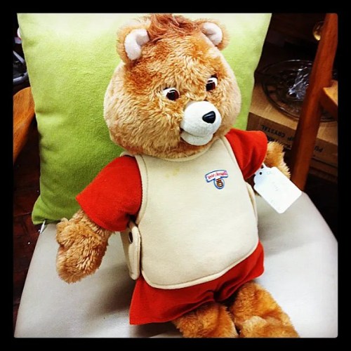 A plush teddy bear wearing a red shirt and beige vest, sitting on a chair next to a green pillow.
