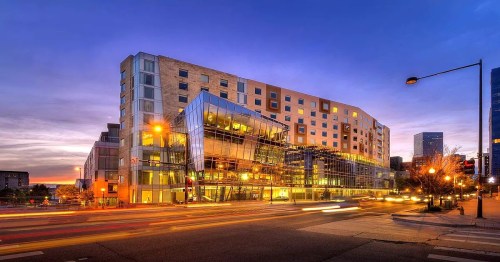 Modern building with glass facade, illuminated at dusk, surrounded by city streets and traffic.