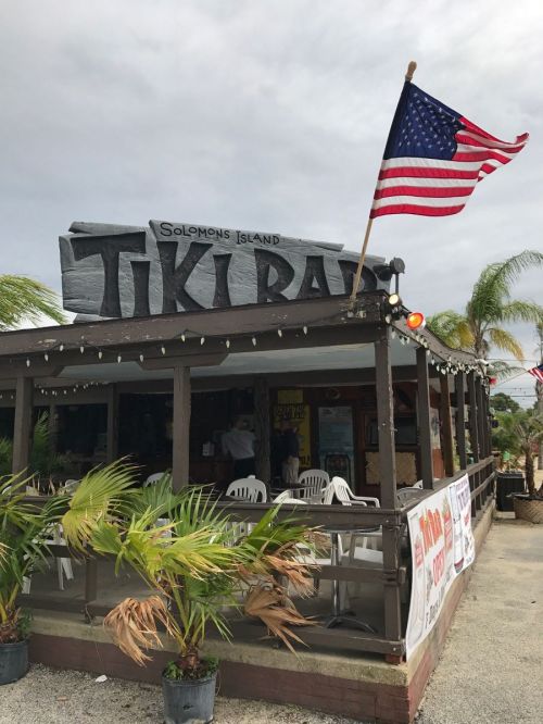 A tiki bar with a sign reading "Tiki Bar," featuring an American flag and palm trees in the foreground.