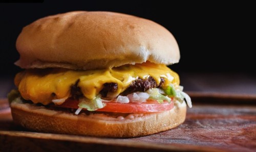A close-up of a cheeseburger with melted cheese, lettuce, tomato, and onions on a soft bun, resting on a wooden surface.