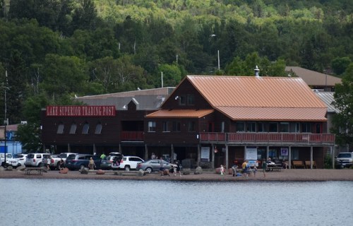 A lakeside trading post with a copper roof, surrounded by trees and people enjoying the beach area.