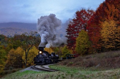A vintage steam train chugs along a curved track surrounded by vibrant autumn foliage.