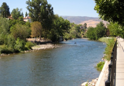 A serene river flows alongside a tree-lined path, with greenery and distant mountains in the background.