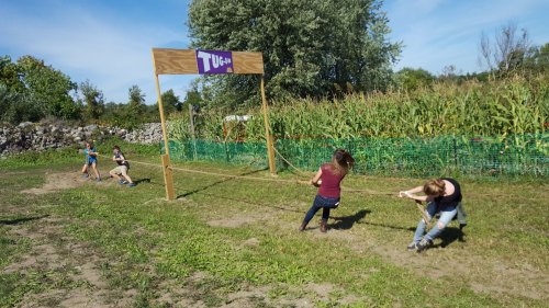 A tug-of-war game in a field, with participants pulling a rope under a sign that reads "Tug." Green cornfield in the background.