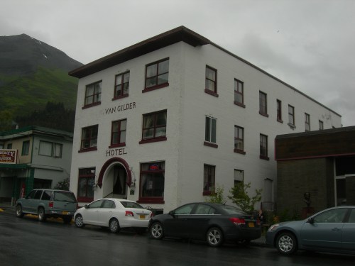 A white three-story hotel with red trim, surrounded by parked cars and mountains in the background on a cloudy day.