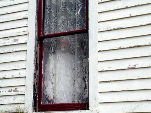 A close-up of a vintage window with lace curtains, set against a weathered white wooden wall.