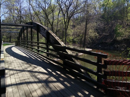 A wooden bridge arches over a calm stream, surrounded by trees and greenery on a sunny day.