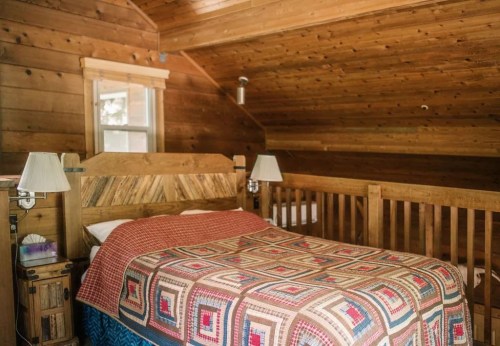 Cozy wooden bedroom with a quilted bedspread, two lamps, and a warm, rustic atmosphere.