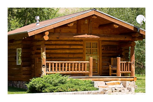 A rustic log cabin with a porch, surrounded by greenery and featuring wooden accents and a satellite dish.