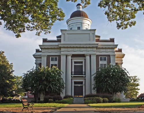Historic building with a dome, large columns, and lush greenery in front, set against a clear sky.