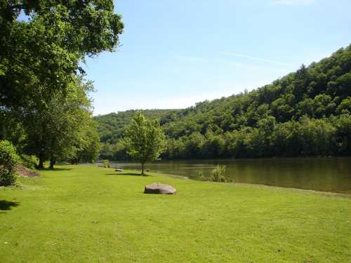 A serene riverbank scene with lush green grass, trees, and a calm river surrounded by hills under a clear blue sky.