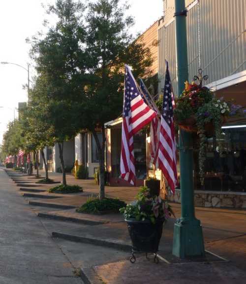 Main street lined with trees and American flags, featuring flower baskets and shops in the background during golden hour.