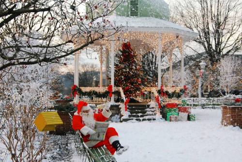 Santa Claus sits on a bench in a snowy park, reading a newspaper near a decorated gazebo and Christmas tree.
