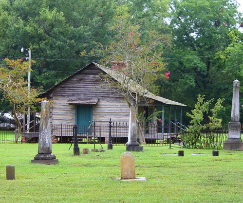 A rustic wooden house surrounded by a cemetery with various gravestones and lush greenery in the background.