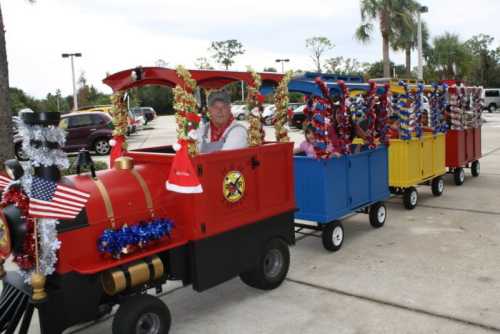 A festive miniature train decorated with tinsel and flags, driven by a man in a red vest, parked in a lot.