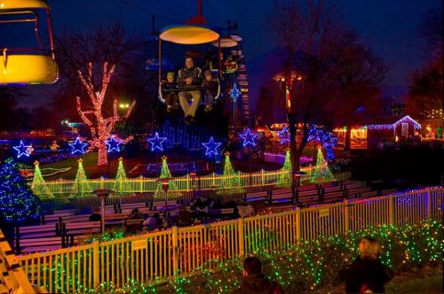 A person rides a chairlift over a festive display of colorful lights and decorated trees at night.